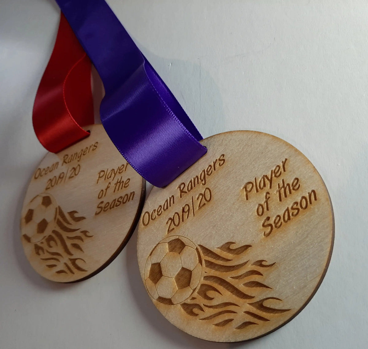 Two round wooden medals hanging against a wall, each laser-engraved with a flaming soccer ball graphic and the text “Ocean Rangers 2019/20 Player of the Season,” suspended by one red and one purple ribbon.