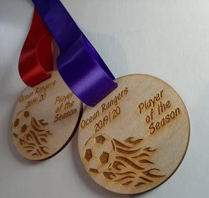Two round wooden medals hanging against a wall, each laser-engraved with a flaming soccer ball graphic and the text “Ocean Rangers 2019/20 Player of the Season,” suspended by one red and one purple ribbon.