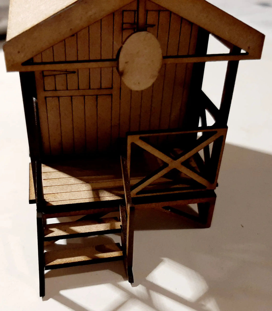 A 1:43 scale model of a beach hut made of wood, featuring detailed construction including a thatched roof and a deck area with chairs.