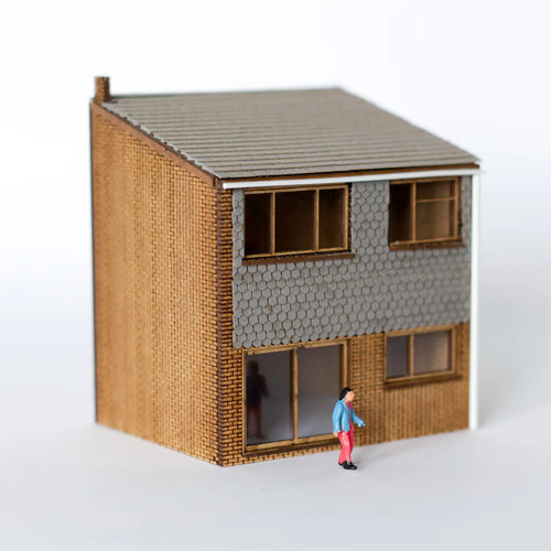 Photograph of a completed Rail Model Terraced House kit in 4 mm scale—a two-storey 1960s-style terrace featuring brick lower walls, grey hexagonal-tile upper cladding, shallow-pitched roof, and sliding patio doors, with a small 4 mm scale figure posed out front for size reference.